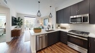 Located in the vibrant Civita community of San Diego, this single-floor residence offers a thoughtfully designed open-concept layout with contemporary finishes throughout. The kitchen is a standout feature, showcasing dark espresso cabinetry that extends to the ceiling, a quartz waterfall peninsula with bar seating, and a full suite of stainless steel appliances including a gas range, microwave, dishwasher, and French door refrigerator. Geometric pendant lighting adds a modern touch, while rich hardwood-style flooring flows seamlessly through the main living areas. The living room is bright and inviting, with direct access to a private balcony that extends the living space outdoors. The bedroom is generously proportioned, featuring large windows that frame views of the surrounding community and fill the room with natural light. A bold teal accent wall and plush carpeting contribute to a warm, stylish atmosphere. The bathroom includes a full soaking tub and shower combination with clean white subway tile surround. A dedicated hallway laundry closet houses a stacked washer and dryer, adding everyday convenience. The overall design balances modern aesthetics with functional living, creating a comfortable and well-appointed home.