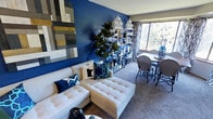 This thoughtfully designed single-floor space makes a strong visual impression through its bold use of color and curated décor. The living area serves as the centerpiece, anchored by a deep royal blue accent wall that creates a dramatic backdrop for a plush white sectional sofa adorned with navy and teal throw pillows. A large tufted ottoman doubles as both a coffee table and additional seating, adding a touch of elegance and practicality to the space.

A striking reclaimed wood wall panel in a geometric patchwork of muted tones provides artistic contrast against the vivid blue wall, lending texture and visual depth to the room. Open metal shelving units line the wall between the living and dining areas, displaying an artful arrangement of white lanterns, decorative objects, and lush indoor greenery, including a statement fiddle-leaf fig tree.

The dining area sits adjacent to a large window that invites generous natural light, illuminating the space with a warm, welcoming glow. A round bistro-style table paired with woven rattan chairs adds a relaxed, café-inspired charm. Bold patterned curtains frame the window, tying the blue-and-white color palette together throughout the space. The overall ambiance is vibrant yet cohesive, blending contemporary design with eclectic, personalized touches.