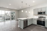 This thoughtfully designed single-floor unit offers a clean, contemporary aesthetic throughout its compact yet efficient layout. The open-concept kitchen and living area serves as the heart of the space, where grey shaker-style cabinetry, white quartz countertops, and a classic subway tile backsplash combine for a polished, modern look. A peninsula island with an undermount sink and pendant lighting above creates a natural gathering point. The kitchen is fully equipped with stainless steel appliances, including a refrigerator, range, built-in microwave, and dishwasher. Warm wood-tone flooring flows throughout the main living area, which is brightened by recessed lighting and a sliding glass door leading to a private balcony with urban views.

The bedroom features large floor-to-ceiling sliding doors that flood the room with natural light and provide direct balcony access, while light hardwood flooring adds a fresh, airy feel. A separate carpeted room offers additional flexible space, finished with neutral tones and recessed ceiling vents. The bathroom is elegantly appointed with a grey vanity, white quartz countertop, a large mirror with contemporary lighting, and a glass-enclosed walk-in shower adorned with veined marble-style tile, delivering a spa-like atmosphere.