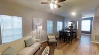 This single-floor space showcases an open-concept layout that seamlessly connects the living, dining, and kitchen areas into one cohesive environment. The vaulted ceiling adds a sense of vertical spaciousness, enhanced by a ceiling fan with integrated lighting that serves both functional and aesthetic purposes.

The living area features warm hardwood-style flooring throughout, complemented by neutral gray walls that create a clean, modern backdrop. Natural light filters in through multiple windows fitted with white horizontal blinds, lending the space a bright and airy feel.

A comfortable seating arrangement anchors the living space, while a natural fiber area rug adds texture underfoot. The dining area accommodates a dark-toned table with matching cross-back chairs, positioned conveniently adjacent to the kitchen. The kitchen, visible in the background, features dark countertops and modern appliances, including a stainless steel refrigerator, accessible via a raised bar-height counter that creates a natural transition between spaces.

Overall, the space presents a well-organized, open floor plan with a cohesive neutral color palette, combining practicality with a relaxed, contemporary aesthetic.