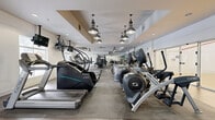 Located at 6005 State Bridge Rd in Johns Creek, GA, this well-appointed fitness center offers a comprehensive, single-level exercise facility designed to support a wide range of workout routines. The space is generously proportioned, providing ample room for both cardio and strength training without feeling crowded.

The cardio zone is lined with a robust selection of equipment including treadmills, elliptical trainers, stair climbers, stationary bikes, and rowing machines from brands such as True, Cybex, and Precor. Overhead, a row of distinctive industrial-style pendant lights runs the length of the space, contributing a modern aesthetic against the clean white ceilings and neutral-toned walls.

The strength training area features power racks, adjustable benches, free weights, barbells, resistance machines, and stability balls, all set on durable rubberized flooring. Full-length mirrors line the walls, enhancing both the sense of space and functional use during workouts. Mounted flat-screen televisions are positioned throughout for convenience.

The flooring transitions between rubberized tiles in the strength zones and wood-look planks in the cardio and transitional areas. Large windows along exterior walls allow abundant natural light to fill the space, offering views of the surrounding community. A standout feature is the adjacent racquetball court, visible through floor-to-ceiling glass panels, adding a unique athletic amenity to this well-equipped facility.