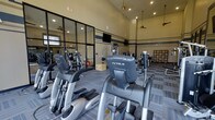 This single-floor fitness center offers a well-appointed workout facility designed to support a full range of exercise routines. The space is characterized by soaring vaulted ceilings that lend an open, airy atmosphere, complemented by ceiling fans that promote airflow throughout. Recessed lighting illuminates the room evenly, creating a bright and energizing environment.

The cardio section is prominently equipped with multiple True-brand elliptical trainers and additional aerobic machines, arranged to allow comfortable spacing between users. A dedicated strength training area features a variety of resistance machines, providing options for both upper and lower body workouts. A mirrored glass-partitioned section along one wall expands the visual depth of the space while allowing exercisers to monitor their form.

A wall-mounted flat-screen television is positioned for easy viewing during workouts. The flooring consists of patterned carpet tiles that define workout zones while providing comfort underfoot. Along one wall, a set of storage lockers offers convenient personal storage for gym users. Decorative horizontal striping along the upper walls adds a structured, purposeful aesthetic to the space. Overall, the fitness center is thoughtfully laid out to maximize functionality while maintaining a clean, professional appearance.