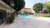 This residential complex centers around a generously sized outdoor swimming pool that serves as the focal point of the community. The pool area is finished with classic brick-edged coping and is surrounded by a spacious concrete deck furnished with multiple lounge chairs, ideal for relaxing under open skies. An adjacent spa provides an additional amenity for residents. Potted palm trees and mature shade trees are thoughtfully positioned throughout the courtyard, adding a lush, California-inspired character to the outdoor environment.

The surrounding buildings feature stucco exteriors in warm earth tones, accented by dark metal balcony railings that line the upper levels. These balconies overlook the pool and courtyard area, creating a sense of community and open connectivity between indoor and outdoor spaces. Ground-level access to the various sections of the complex is managed through secured black iron gates, ensuring privacy and safety for residents.

Landscaped green lawns, ornamental shrubs, and garden beds are interspersed throughout the grounds, softening the hardscape and contributing to a pleasant, resort-like atmosphere. Stairwells and walkways connect the levels, with mature trees providing natural shade. The overall ambiance is that of a comfortable, well-kept community with a strong emphasis on outdoor living.