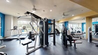 This fitness center offers a thoroughly equipped workout space designed to support a wide range of exercise routines. The interior is defined by a bold color palette, with striking blue accent walls and yellow ceiling details that lend the space a dynamic, energetic atmosphere. Large windows line the perimeter, flooding the floor with natural light and providing views of the surrounding outdoor greenery.

The space is organized to accommodate both cardiovascular training and strength work. The cardio section features an array of machines including treadmills, ellipticals, stair climbers, and stationary bikes, many equipped with digital displays. The strength training area is equally well-appointed, with a multi-station cable machine, a Smith machine with a full bench and weight plates, a dedicated free weight station with a complete kettlebell and dumbbell rack, and additional selectorized weight machines.

Ceiling fans and recessed lighting ensure comfort and visibility throughout the space. Full-length mirrors are integrated into portions of the walls, visually expanding the room and supporting proper form during workouts. The durable rubber and concrete flooring is practical and easy to maintain. Multiple entry and exit points, including a direct exterior door, add to the convenience and accessibility of this fitness facility.