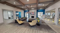 Located at 2650 Western Center Blvd in Fort Worth, TX, this single-story community clubhouse and leasing center offers a thoughtfully designed space that blends modern aesthetics with functional amenities. The centerpiece of the facility is a spacious lounge area distinguished by soaring coffered ceilings with exposed wood beams, a striking wrought-iron chandelier, and light wood-look flooring throughout. A tile-surround fireplace anchors one wall beneath a mounted flat-screen TV, while comfortable gray sofas and accent chairs in warm yellow tones create inviting seating arrangements. Bold teal accent walls punctuate the bright, white interior, lending the space a vibrant and contemporary character. The entry foyer features additional teal upholstered seating and transitions seamlessly into a staffed reception desk area highlighted by a vivid blue backdrop. A well-appointed kitchenette/refreshment bar includes dark countertops, white cabinetry, floating shelves adorned with colorful décor, a coffee station, and a stainless steel dishwasher. A dedicated business center offers a communal high-top worktable with computer stations set against a deep blue accent wall, providing a productive workspace. Abundant natural light filters in through multiple windows, and glass-panel doors open to outdoor views, enhancing the overall sense of openness throughout the facility.