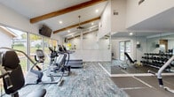 This fitness center offers a thoughtfully designed, single-level layout that accommodates a comprehensive range of workout equipment across distinct activity zones. The space is defined architecturally by soaring vaulted ceilings accented with prominent exposed wood beams, lending a warm, rustic contrast to the otherwise clean, white-walled interior. Recessed lighting and a ceiling fan complement the natural light that streams in through multiple windows and glass entry doors, creating a bright and energizing atmosphere.

The cardio zone houses a row of treadmills, ellipticals, and a stationary bike, all positioned along windowed walls that provide views of the surrounding outdoor greenery. Wall-mounted televisions are installed throughout for entertainment during workouts. A large floor-to-ceiling mirror spans one section of the space, visually expanding the room and supporting stretching and functional training activities.

A dedicated strength training area features several selectorized cable and weight machines, including leg press and pulldown equipment. One wall in this zone is finished with warm wood-toned horizontal planking, adding a distinct design element beneath a mounted flat-screen television. The flooring throughout combines patterned carpet tiles with rubberized mat sections in the free-weight area, providing both comfort and functionality across workout zones.