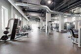This expansive fitness facility offers a comprehensive, well-equipped workout environment spread across a single, open floor plan. The industrial aesthetic is defined by soaring ceilings with exposed ductwork, dark-painted structural elements, and bold white support columns that frame the space throughout. Durable rubber flooring covers the main training areas, providing a safe and functional surface for a wide range of exercises.

The main floor is organized into distinct training zones. A robust strength training section houses a broad selection of selectorized machines, free weights, benches, and cable systems. The cardio area features rowing machines and cycling bikes. A dedicated functional training zone includes a multi-purpose rig with suspension trainers, battle ropes, kettlebells, medicine balls, and a marked turf sprint lane. Natural light filters in through high clerestory windows, complementing the overhead track lighting throughout.

Separate studio rooms with warm hardwood flooring offer versatile spaces for group fitness classes, yoga, stretching, or personal training sessions. These rooms feature clean white walls, wall-mounted displays, and LED strip lighting, providing a quieter, focused environment distinct from the main floor. The facility's scale and variety of equipment make it a fully self-contained training destination suited for diverse fitness needs.