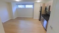 This single-floor space offers a clean, functional layout centered around an open-plan living and kitchen area. The light hardwood flooring runs throughout, lending warmth and visual continuity to the interior. Natural light filters in through a horizontal window positioned near the ceiling, characteristic of a below-grade or garden-level setting, giving the space an airy quality despite its intimate scale.

The kitchen is efficiently designed and positioned along one wall, featuring a full suite of stainless steel appliances, including a gas range with oven, refrigerator, and microwave. Light wood cabinetry complements the flooring and contributes to the overall bright, neutral palette of the space. The open connection between the kitchen and living area maximizes the sense of space, making the layout versatile and well-suited for everyday living.

The walls are finished in a crisp off-white tone, and clean white baseboards frame the perimeter of the room. Flush ceiling lighting ensures even illumination throughout. The overall ambiance is modern, minimal, and move-in ready, with a practical design that makes efficient use of the available footprint while maintaining a tidy and welcoming atmosphere.