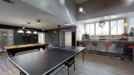 This single-floor entertainment space is designed with leisure and social interaction in mind. The game room serves as the centerpiece, featuring a full-sized pool table illuminated by a classic pendant light fixture, alongside a ping pong table positioned prominently in the open floor plan. The space benefits from soaring ceilings that create an airy, expansive atmosphere, with natural light filtering in through windows on multiple walls.

The interior is finished in a contemporary style, with dark gray walls that contribute to a sleek, modern aesthetic. Light wood-toned flooring provides a warm contrast to the cooler tones of the walls and furnishings. A wet bar or kitchenette area is visible toward the rear of the space, complete with bar-height seating, pendant lighting, and counter space — ideal for entertaining guests. Recessed and decorative ceiling fixtures provide additional ambient lighting throughout.

A doorway connects to an adjacent area, suggesting further utility within the layout. Overall, the space is thoughtfully arranged to accommodate multiple recreational activities simultaneously, making it well-suited as a community or private entertainment venue.