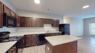 This single-floor unit is designed with a functional, open layout that connects the living and kitchen areas seamlessly. The kitchen is a standout feature, showcasing rich dark wood cabinetry with sleek hardware, light granite countertops, and a full suite of black appliances including a range, over-the-range microwave, dishwasher, and refrigerator. A central island with a matching granite surface provides additional workspace and serves as a natural focal point. Light wood-style flooring runs throughout the main living areas, contributing to a clean, contemporary aesthetic.

The living area is generously proportioned, illuminated by recessed lighting and a ceiling fan, and benefits from natural light through blinds-fitted windows. Neutral gray walls create a versatile backdrop throughout the unit.

The space includes at least two bathrooms: one features a glass-enclosed walk-in shower with white subway tile surround and granite vanity, while the second offers a tub/shower combination with matching finishes and tile flooring. A spacious walk-in closet with wire shelving provides ample storage. Overall, the unit presents a cohesive, modern finish package throughout.