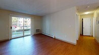 This 1-bedroom, 1-bathroom unit is thoughtfully laid out across a single floor, offering a clean and functional living environment. The living area features warm hardwood flooring and neutral white walls that create a bright, open feel. A sliding glass door allows natural light to pour in and provides direct views of the surrounding outdoor greenery. Baseboard heating runs throughout the main living spaces, ensuring year-round comfort. A wall-mounted air conditioning unit adds climate flexibility.

The kitchen is efficiently designed in a U-shaped layout, outfitted with white cabinetry offering generous storage, light-toned countertops, and a full suite of white appliances including an electric range, built-in microwave, dishwasher, and refrigerator. Hardwood flooring continues seamlessly from the living area into the kitchen, which is illuminated by overhead lighting. A breakfast bar provides a convenient transition between the kitchen and dining area, which is accented by a classic chandelier.

The bedroom is carpeted for comfort and features multiple windows that invite abundant natural light and neighborhood views. A closet with a rod and shelving provides practical storage. The full bathroom includes a tub/shower combination, toilet, and a wall-mounted cabinet for additional storage. Overall, the unit presents a clean, move-in-ready aesthetic with a practical and comfortable layout.
