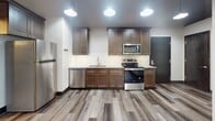 This single-floor space offers a compelling mix of industrial heritage and modern renovation, creating a distinctive urban loft aesthetic throughout. The fully updated kitchen serves as a standout feature, showcasing rich dark-stained cabinetry, granite countertops, a classic white subway tile backsplash, and a full suite of stainless steel appliances including a refrigerator, electric range, microwave, and dishwasher. Pendant lighting and wide-plank wood-style flooring complete the kitchen's polished, contemporary look.

The space includes multiple rooms that reflect a variety of finish levels, highlighting the building's evolving character. Some rooms feature the same warm wood-style flooring found in the kitchen, while others retain their original polished or painted concrete floors — a nod to the structure's industrial roots. Exposed wooden ceiling beams run throughout, adding texture and architectural interest overhead. Windows fitted with roller shades allow for natural light while maintaining privacy.

The bathrooms are clean and modern, equipped with walk-in showers featuring white panel surrounds, linear drains, built-in shelving niches, and granite-topped vanities with dark cabinetry that mirrors the kitchen's design language. The overall ambiance balances raw, character-rich elements with thoughtfully updated finishes.