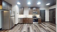 This single-floor space offers a compelling mix of industrial heritage and modern renovation, creating a distinctive urban loft aesthetic throughout. The fully updated kitchen serves as a standout feature, showcasing rich dark-stained cabinetry, granite countertops, a classic white subway tile backsplash, and a full suite of stainless steel appliances including a refrigerator, electric range, microwave, and dishwasher. Pendant lighting and wide-plank wood-style flooring complete the kitchen's polished, contemporary look.

The space includes multiple rooms that reflect a variety of finish levels, highlighting the building's evolving character. Some rooms feature the same warm wood-style flooring found in the kitchen, while others retain their original polished or painted concrete floors — a nod to the structure's industrial roots. Exposed wooden ceiling beams run throughout, adding texture and architectural interest overhead. Windows fitted with roller shades allow for natural light while maintaining privacy.

The bathrooms are clean and modern, equipped with walk-in showers featuring white panel surrounds, linear drains, built-in shelving niches, and granite-topped vanities with dark cabinetry that mirrors the kitchen's design language. The overall ambiance balances raw, character-rich elements with thoughtfully updated finishes.