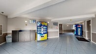 Located at 9555 128 Street in Surrey, BC, this building lobby serves as the main entry point for residents and visitors. The space is composed of a single-floor hallway layout that provides a functional and practical welcome area. Light blue ceramic floor tiles with a geometric diagonal grout pattern extend throughout, giving the space a clean, uniform appearance. The walls are finished in a two-tone grey scheme, divided by a white chair rail, complemented by a textured ceiling with crown moulding detailing. Wall-mounted sconce lighting provides ambient illumination throughout the corridor. A key feature of the lobby is the bank of individual resident mailboxes mounted along one wall, offering convenient mail access. Beverage vending machines — including a Pepsi unit — are positioned within the hallway, adding practical amenity for building occupants. A reception or package counter area is also present. The main entrance features a glass sliding door that floods the space with natural light and provides a clear view of the surrounding outdoor greenery and parking area. Entry mats branded with "Mainstreet" are placed at access points. Baseboard heating runs along the perimeter walls, ensuring comfort year-round. The overall ambiance is utilitarian and well-maintained, designed to efficiently serve the daily needs of building residents.