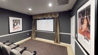 This private home theater is designed to deliver an authentic cinematic experience from the comfort of home. The room is finished in deep charcoal-gray walls accented by white chair-rail molding that runs the perimeter, creating a sleek, theater-inspired aesthetic. Recessed ceiling lighting and wall-mounted sconces provide adjustable ambient illumination throughout the space.

At the front of the room, a large projection screen is framed by elegant draped gold curtains with a matching decorative valance, lending a classic movie-house feel. A ceiling-mounted projector is centrally positioned to deliver a crisp, large-format picture.

The seating area is arranged in two tiered rows of stadium-style theater seats, each upholstered in gray fabric with black armrests and built-in cup holders, ensuring comfort during any screening. The raised rear platform provides unobstructed sightlines from every seat. Plush dark carpeting covers both the floor and the stepped risers, contributing to the room's acoustically controlled environment.

The side walls are adorned with framed classic film posters — including Star Wars, Titanic, and Casablanca — adding a curated, cinephile character to the space. An AV equipment cabinet is neatly integrated into the seating area. The room is completed by a dedicated entry door, making this a fully self-contained, purpose-built private cinema.