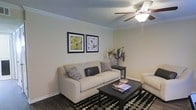 This single-floor space offers a comfortable and well-appointed living environment. The living room serves as the focal point of the home, showcasing a warm and inviting atmosphere with a neutral color palette on the walls that complements the dark hardwood-style flooring throughout. Crown molding adds a refined architectural detail along the ceiling perimeter, lending a classic and polished character to the room. A ceiling fan with a built-in light fixture is centrally positioned, providing both airflow and ambient lighting. The textured ceiling adds subtle visual depth overhead. The space accommodates a generously sized sofa alongside additional seating, centered around a dark coffee table and anchored by a bold black-and-white patterned area rug. Decorative framed artwork adorns the walls, contributing to the curated aesthetic of the room. A side table with a floral arrangement adds a natural, decorative touch. The overall ambiance of the space is cozy yet tasteful, with thoughtful design details that create a functional and welcoming living environment suited for everyday comfort.