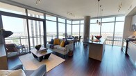 This sophisticated high-rise residence offers an impressive open-plan layout that seamlessly integrates the living, dining, and kitchen areas into one cohesive, light-filled space. Floor-to-ceiling wraparound windows flood the interior with abundant natural light while framing breathtaking panoramic views of the city skyline and surrounding landscape, creating an immediate sense of grandeur and elevation.

The rich, dark hardwood flooring runs throughout, providing a warm and elegant foundation that contrasts beautifully with the bright, airy atmosphere created by the expansive glazing. A striking exposed concrete column anchors the space architecturally, adding an industrial character that complements the contemporary design aesthetic.

The living area is centered around a comfortable sofa accented with bold navy and yellow throw pillows, paired with a low-profile wooden coffee table. Adjacent to it, the dining area accommodates a full table and chairs, benefiting directly from the dramatic window views. The kitchen features a sleek dark island with bar seating, pendant lighting overhead, and well-appointed counter space, making it both functional and visually refined. Track lighting along the ceiling provides additional illumination throughout. The overall ambiance is modern, elevated, and urbane, with a strong emphasis on views, natural light, and high-end finishes.