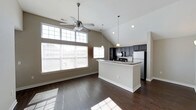 Located at 11289 E Hamilton Crest Blvd in Fishers, Indiana, this single-floor home offers a thoughtfully designed open-concept layout that blends comfort and functionality. The main living area is defined by soaring vaulted ceilings that amplify the sense of space, while rich dark hardwood floors run throughout, adding warmth and elegance. A ceiling fan with integrated lighting serves the living area, which benefits from an abundance of natural light streaming through a large multi-pane window bank along the exterior wall. The open kitchen is a standout feature, showcasing dark cabinetry, stone countertops, and stainless steel appliances including a refrigerator and range. A central kitchen island with a contrasting light finish provides additional prep and seating space, while pendant lighting above adds a modern, polished touch. The seamless flow between the kitchen and living area makes this home well-suited for both everyday living and entertaining. Neutral-toned walls throughout complement the overall color palette, creating a cohesive and inviting atmosphere. The home's single-floor design ensures easy accessibility and a streamlined living experience in a well-established Fishers community.
