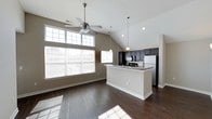 Located at 11289 E Hamilton Crest Blvd in Fishers, Indiana, this single-floor home offers a thoughtfully designed open-concept layout that blends comfort and functionality. The main living area is defined by soaring vaulted ceilings that amplify the sense of space, while rich dark hardwood floors run throughout, adding warmth and elegance. A ceiling fan with integrated lighting serves the living area, which benefits from an abundance of natural light streaming through a large multi-pane window bank along the exterior wall. The open kitchen is a standout feature, showcasing dark cabinetry, stone countertops, and stainless steel appliances including a refrigerator and range. A central kitchen island with a contrasting light finish provides additional prep and seating space, while pendant lighting above adds a modern, polished touch. The seamless flow between the kitchen and living area makes this home well-suited for both everyday living and entertaining. Neutral-toned walls throughout complement the overall color palette, creating a cohesive and inviting atmosphere. The home's single-floor design ensures easy accessibility and a streamlined living experience in a well-established Fishers community.