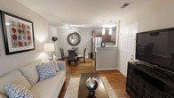 This single-floor residence offers an open-concept layout that seamlessly connects the living, dining, and kitchen areas into one cohesive and functional space. The living area is anchored by a light gray sofa, complemented by a plush area rug and a sleek rectangular coffee table, creating an inviting and comfortable atmosphere. Warm hardwood-style flooring runs throughout, adding continuity and a sense of warmth to the entire space.

The dining area is positioned adjacent to the living room and features a round glass-top table surrounded by upholstered chairs, with a decorative circular mirror serving as a stylish focal point on the wall behind it. This arrangement makes the space feel open while maintaining a defined dining zone.

The kitchen is accessible via a half-wall peninsula that provides both separation and connection to the main living area. It features medium-toned wood cabinetry, a stainless steel refrigerator, and pendant lighting that adds a modern touch overhead. The layout is efficient and well-organized, making it practical for everyday use.

The space is finished with neutral white walls, recessed and track lighting, and clean white trim throughout, contributing to a bright, modern, and well-maintained overall ambiance.
