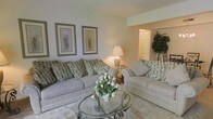 This single-floor residence offers a thoughtfully designed layout that balances comfort and classic style. The living area serves as the heart of the space, featuring plush, neutral-toned sofas accented with patterned throw pillows that add subtle texture and visual interest. A round glass-top coffee table with an ornate metal base anchors the seating arrangement, while a decorative floral centerpiece brings a touch of warmth and color to the room. Soft, warm lighting from a classic table lamp enhances the cozy atmosphere, complementing the light-colored carpeted flooring that runs throughout. Framed botanical artwork on the walls adds a refined decorative touch, contributing to the space's understated elegance. The open layout flows naturally toward adjacent areas, with a glimpse of a doorway leading to connected rooms, suggesting a well-connected and functional floor plan. A lush indoor plant near the entryway adds a natural, lively element to the overall design. The consistent use of soft beige and warm neutral tones throughout creates a cohesive, serene environment that feels both welcoming and timeless.