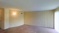 This single-floor space is defined by its striking white-painted brick walls, which wrap around the interior and serve as the room's most prominent architectural feature. The painted brick adds texture and visual interest while maintaining a clean, bright aesthetic that reflects light throughout the space. The flooring consists of wall-to-wall carpeting in a neutral mauve-gray tone, contributing to a warm and comfortable atmosphere underfoot. The smooth white ceiling provides a clean contrast to the rugged texture of the brick below. Wall-mounted sconce lighting is installed directly onto the brick surface, offering a practical and decorative lighting solution that enhances the room's cozy ambiance. Electrical outlets are positioned at standard intervals along the brick walls, ensuring functional usability throughout the space. The open layout provides versatile room for a variety of furniture arrangements and uses. Overall, the combination of textured brick walls, neutral carpeting, and warm accent lighting creates a casual, inviting environment with a distinct character rooted in classic masonry construction.