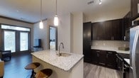 This single-floor residence showcases a thoughtfully designed open-concept layout that seamlessly connects the kitchen and living areas. The kitchen serves as a central hub, anchored by a large granite-topped island with an undermount sink and a brushed nickel pull-down faucet. Two bar stools tuck neatly beneath the island's overhang, creating a casual dining and gathering space. Pendant lights with sleek cylindrical shades hang above the island, providing focused task lighting with a contemporary aesthetic.

Dark espresso-toned cabinetry lines the kitchen walls, offering generous storage with clean bar-pull hardware. A classic white subway tile backsplash provides a crisp contrast against the darker cabinetry. Light wood-look flooring runs throughout, adding warmth while complementing the neutral color palette of the walls. Recessed track lighting in the ceiling supplements the pendant fixtures, ensuring the space is well-lit throughout.

A glimpse into the adjacent bedroom reveals a bright, comfortable room with a ceiling fan and ample natural light, reflecting the cohesive and modern character carried throughout the entire residence.