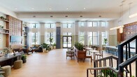 Located at 1104 Avondale Ave SE in Atlanta, Georgia, this single-floor space offers a generously sized, open layout that makes a strong architectural statement. Light-toned hardwood flooring runs throughout, anchoring the airy, well-lit interior and contributing to a warm, cohesive aesthetic.

The living room is the centerpiece of the space, commanding attention with soaring double-height ceilings and floor-to-ceiling windows that flood the interior with natural light and offer views of the surrounding neighborhood. Sheer white curtains frame the oversized windows, softening the light while maintaining an open, expansive feel.

A standout design feature is the custom-built walnut media wall, which combines a sculptural, circular-patterned panel above with rich cabinetry below. Flanking open shelving provides display and storage space, accessorized with plants, books, and décor that add color and life to the wall. Circular wall-mounted sconces on either side provide warm accent lighting.

The broader space incorporates a multi-zone layout, including a lounge area with blue upholstered seating, a staircase with black iron railings, and lush greenery throughout. Tall structural columns with wainscoting painted in a muted blue-gray tone add classical character. The overall ambiance is sophisticated yet approachable — a blend of modern design sensibility and warm, residential comfort.