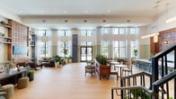 Located at 1104 Avondale Ave SE in Atlanta, Georgia, this single-floor space offers a generously sized, open layout that makes a strong architectural statement. Light-toned hardwood flooring runs throughout, anchoring the airy, well-lit interior and contributing to a warm, cohesive aesthetic.

The living room is the centerpiece of the space, commanding attention with soaring double-height ceilings and floor-to-ceiling windows that flood the interior with natural light and offer views of the surrounding neighborhood. Sheer white curtains frame the oversized windows, softening the light while maintaining an open, expansive feel.

A standout design feature is the custom-built walnut media wall, which combines a sculptural, circular-patterned panel above with rich cabinetry below. Flanking open shelving provides display and storage space, accessorized with plants, books, and décor that add color and life to the wall. Circular wall-mounted sconces on either side provide warm accent lighting.

The broader space incorporates a multi-zone layout, including a lounge area with blue upholstered seating, a staircase with black iron railings, and lush greenery throughout. Tall structural columns with wainscoting painted in a muted blue-gray tone add classical character. The overall ambiance is sophisticated yet approachable — a blend of modern design sensibility and warm, residential comfort.