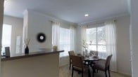 This single-story space showcases a thoughtfully designed interior that balances comfort and style. The dining area is a standout feature, bathed in natural light streaming through multiple large windows dressed with sheer white curtains and horizontal blinds. The windows offer pleasant outdoor views of surrounding greenery, enhancing the airy atmosphere of the room. Crown molding lines the ceiling throughout, adding a refined architectural detail that elevates the overall aesthetic. Recessed lighting complements the natural light, ensuring the space remains well-illuminated. The dining area accommodates a round dark-wood table surrounded by upholstered chairs, creating an inviting setting for meals and gatherings. A decorative starburst wall mirror adds a stylish focal point to the neutral-toned walls. A partial counter or breakfast bar is visible, suggesting an open connection to an adjacent kitchen area, promoting a seamless flow between spaces. The light-colored walls, neutral carpeting, and clean lines contribute to a cohesive, modern interior design. Overall, the space presents a warm, welcoming ambiance enhanced by abundant natural light and tasteful finishes.