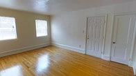 This single-floor space presents a straightforward and functional layout with a clean, neutral aesthetic throughout. The main living area immediately draws attention with its warm-toned hardwood flooring, which runs continuously and reflects natural light beautifully, adding a sense of warmth and openness to the room. The walls are finished in a crisp, off-white tone, providing a bright and versatile backdrop that complements a variety of furnishing styles. A window fitted with horizontal blinds allows natural light to filter into the space while maintaining privacy, contributing to the airy feel of the interior. A classic six-panel white door with a brass lever handle serves as the entry point, adding a touch of traditional character to the overall design. White painted baseboards frame the perimeter of the room, providing a neat, finished look that ties the space together. The layout is open and uncluttered, making it highly adaptable for a range of uses. Overall, the space combines practical design elements with warm, natural materials to create a welcoming and functional environment.