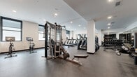 Located at 1229 Baltimore Ave in the heart of Kansas City, MO, this fitness center offers a generously sized, single-floor exercise room designed to support a comprehensive workout experience. The space features a clean, modern aesthetic with white walls, structural columns, and a dark rubber-composite floor that provides both durability and comfort underfoot. Recessed lighting throughout keeps the room bright and well-illuminated.

The exercise room is thoughtfully equipped with a full lineup of cardio machines, including multiple treadmills and ellipticals, as well as a multi-station cable machine, weight bench, rowing machines, and a dedicated dumbbell rack. Spin bikes with integrated screens add a modern, tech-enabled cardio option. A large floor-to-ceiling mirror spans one wall, visually expanding the space and supporting proper form during workouts.

A standout feature is the bold, large-scale mural depicting jazz musicians in a warm palette of oranges and reds — a nod to Kansas City's rich musical heritage — which serves as a vibrant focal point anchored by a mounted flat-screen television. Natural light filters in through multiple windows along the perimeter walls, enhancing the open, energizing atmosphere. The overall design balances functionality with character, making this fitness center both practical and visually engaging.