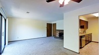 This single-story space presents a straightforward and functional layout centered around a carpeted living area. The room is finished with neutral white walls and gray wall-to-wall carpeting, creating a clean, blank-canvas aesthetic well-suited for personalization. A ceiling fan with a light fixture is mounted overhead, providing both air circulation and ambient lighting to the space.

A standout feature of the living area is the brick fireplace, positioned in the corner of the room. The fireplace is framed with brick detailing and includes a raised hearth with tiled accents, adding a touch of warmth and character to the otherwise understated interior. A dark-stained wooden door is visible nearby, providing access to an adjacent room or hallway.

The space benefits from a practical design with smooth, white ceilings and standard baseboards throughout. A wall-mounted thermostat or light switch panel is visible near the doorway, and a ceiling vent indicates a central HVAC system. Overall, the space offers a modest, livable environment with classic residential features that provide a solid foundation for comfortable everyday living.