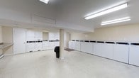 This shared laundry facility offers a clean, functional, and well-organized space designed to accommodate a high volume of laundry needs. The room is finished with smooth, light-toned flooring and warm beige walls that contribute to a bright and welcoming atmosphere. Overhead fluorescent lighting ensures the entire space is well-lit throughout the day.

A long row of top-loading washing machines lines one wall, providing ample capacity for simultaneous use by multiple residents. Opposite the washers, a bank of white stacked dryer units offers equally generous drying capacity, neatly arranged in uniform cabinetry for a tidy appearance. A central structural column divides the open floor plan subtly without impeding the flow of the space.

The layout is thoughtfully designed to allow comfortable movement between machines, with generous open floor space in the center. A recessed skylight or ceiling vent adds to the room's ventilation and airflow. The overall ambiance is practical and efficient, with a no-frills, clean aesthetic that prioritizes ease of use. This laundry room serves as a convenient shared amenity, fully equipped to meet everyday laundry demands in a communal setting.
