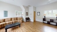 Located in Winston-Salem, NC, this single-story space offers a well-designed layout that balances openness with functionality. The main living area is a standout feature, characterized by vaulted ceilings that add a sense of height and airiness to the room. Warm-toned hardwood flooring runs throughout the primary living spaces, contributing to a cohesive and inviting aesthetic.

The main lounge area is generously sized, furnished with a striped sofa, a leather armchair, and a dark wood coffee table. Large multi-pane windows on multiple walls flood the room with natural light and provide views of the surrounding outdoor environment. A set of glass-paneled double doors offers direct exterior access and further enhances the brightness of the space.

An adjacent area with vaulted architectural detailing provides additional seating space, accented by framed wall art and a classic wooden bench. This section features a charming cottage-style window that frames natural greenery outside.

The space also includes a dedicated laundry room, finished with light tile flooring, a stacked washer and dryer unit, a long folding table, and a large window that keeps the area bright and practical. Overall, the space presents a clean, functional, and comfortable environment suitable for communal use.