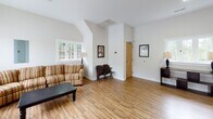 Located in Winston-Salem, NC, this single-story space offers a well-designed layout that balances openness with functionality. The main living area is a standout feature, characterized by vaulted ceilings that add a sense of height and airiness to the room. Warm-toned hardwood flooring runs throughout the primary living spaces, contributing to a cohesive and inviting aesthetic.

The main lounge area is generously sized, furnished with a striped sofa, a leather armchair, and a dark wood coffee table. Large multi-pane windows on multiple walls flood the room with natural light and provide views of the surrounding outdoor environment. A set of glass-paneled double doors offers direct exterior access and further enhances the brightness of the space.

An adjacent area with vaulted architectural detailing provides additional seating space, accented by framed wall art and a classic wooden bench. This section features a charming cottage-style window that frames natural greenery outside.

The space also includes a dedicated laundry room, finished with light tile flooring, a stacked washer and dryer unit, a long folding table, and a large window that keeps the area bright and practical. Overall, the space presents a clean, functional, and comfortable environment suitable for communal use.