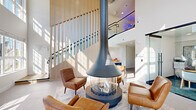 This thoughtfully designed lobby space makes an immediate impression with its soaring double-height ceilings and expansive floor-to-ceiling windows that flood the interior with natural light. The architectural centerpiece is a striking suspended pendant fireplace, encased in a cylindrical glass surround and anchored by a circular base, which commands the living room area and draws the eye upward toward the dramatic vertical volume above. Surrounding the fireplace, a curated seating arrangement of cognac leather armchairs rests atop a patterned area rug, creating an inviting and refined gathering point. The living room's crisp white walls and light-toned tile flooring contrast warmly with the wood-paneled staircase railing, which frames the mezzanine level above — complete with modern metal railings and accent lighting. An open staircase connects to the upper level, reinforcing the sense of vertical openness throughout. The hallway leads to a glass-enclosed leasing office, offering a transparent yet private workspace bathed in natural light. Adjacent to it, a bank of individual mailboxes lines the corridor wall, reflecting the building's functional, resident-focused design. Together, these spaces combine modern aesthetics with practical amenities to create a polished, welcoming communal environment.