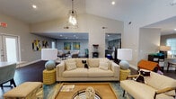 This single-floor community space is designed with an open-concept layout that seamlessly connects multiple functional areas, creating a welcoming and versatile environment. The main lounge area is anchored by a generously sized neutral sofa, complemented by stylish globe-base lamps, a wood-and-metal coffee table, and a patterned area rug in blue and teal tones. Soaring vaulted ceilings with an industrial-style cage pendant light give the space a dramatic sense of height and openness.

A statement wall clad in white shiplap serves as the backdrop for a wall-mounted television, adding a modern farmhouse touch to the lounge. Adjacent to the lounge is a large dining area furnished with a long table and upholstered teal chairs, bathed in natural light from French doors that open to an outdoor area with pool views. Colorful artwork adorns the walls throughout, adding energy and character.

The kitchen area features gray cabinetry, a breakfast bar with teal barstools, and stainless steel appliances including a full-size refrigerator. A popcorn machine adds a fun, recreational touch. A glass-windowed office or leasing area is also accessible from the main corridor, alongside a workstation zone with individual desks. Rich dark hardwood flooring flows throughout, unifying the entire space.