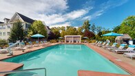 Located at 299 Colt Hwy in Farmington, Connecticut, this outdoor pool area offers a well-appointed retreat designed for relaxation and recreation. The centerpiece is a generously sized swimming pool with crystal-clear turquoise water, framed by a broad brick-paved deck that provides ample space for lounging and socializing. A dedicated hot tub or wading area is accessible at one end of the pool, adding to the versatility of the aquatic amenities.

The deck is furnished with a full complement of cushioned chaise lounges and chairs, many shaded by vibrant blue market umbrellas, creating a comfortable environment for sunbathing or unwinding. A standout architectural feature is the white pergola cabana structure at one end of the deck, offering a shaded gathering spot with additional seating for guests seeking refuge from the sun.

The surrounding landscape features mature deciduous trees and ornamental plantings that provide natural privacy and a lush, park-like atmosphere. The pool area is enclosed by a classic black iron fence, ensuring a secure and defined boundary. Overlooking the space are multi-story buildings with traditional New England-style architecture, characterized by brick and siding exteriors, pitched rooflines, and large windows. A clubhouse-style building with floor-to-ceiling windows adjoins the pool deck, providing seamless indoor-outdoor connectivity and completing this inviting outdoor amenity space.