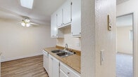Located in San Lorenzo, CA, this one-bedroom, one-bathroom unit offers a practical and well-organized single-floor layout. The combined kitchen and living area forms the heart of the home, with warm wood-look flooring running throughout. The galley-style kitchen is efficiently designed with white cabinetry on both sides, stone-tone countertops, a double-basin sink, and white appliances including a dishwasher, range, and refrigerator — all illuminated by overhead lighting. The living room is open and inviting, featuring a ceiling fan, a wall-mounted heater, and a sliding glass door that fills the space with natural light and provides access to an outdoor area with views of surrounding greenery. The bedroom is a comfortable, well-proportioned room with soft carpeting, a window that brings in natural light, and a dedicated closet for storage. The bathroom is clean and functional, outfitted with a white vanity and cabinet, a mirrored medicine cabinet, wood-look flooring, and a full tub and shower enclosed by sliding glass doors. Neutral, light-toned walls throughout the unit create a bright and cohesive atmosphere, making this a practical and comfortable living space.