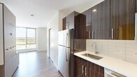 Located on the fourth floor at 199 N Harvard St in Boston's Allston neighborhood, this efficiently designed studio unit offers a well-appointed layout across a single floor. The kitchen is a standout feature, showcasing rich dark wood grain cabinetry with a high-gloss finish, white quartz countertops, a stainless steel undermount sink with a modern arc faucet, white subway tile backsplash, and stainless steel appliances including a full-size refrigerator and range. The open living area is anchored by wide-plank wood-look flooring and a large floor-to-ceiling window that floods the space with natural light and offers elevated views of the surrounding Boston cityscape. A built-in multi-functional furniture unit with integrated shelving, storage compartments, and a fold-out surface maximizes the use of the compact footprint, providing smart organization solutions throughout. The bathroom features a clean, contemporary design with a white vanity, quartz countertop, undermount sink, chrome fixtures, and a backlit vanity mirror that adds a refined touch. Neutral white walls and consistent flooring create a cohesive, modern aesthetic throughout the unit.