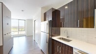 Located on the fourth floor at 199 N Harvard St in Boston's Allston neighborhood, this efficiently designed studio unit offers a well-appointed layout across a single floor. The kitchen is a standout feature, showcasing rich dark wood grain cabinetry with a high-gloss finish, white quartz countertops, a stainless steel undermount sink with a modern arc faucet, white subway tile backsplash, and stainless steel appliances including a full-size refrigerator and range. The open living area is anchored by wide-plank wood-look flooring and a large floor-to-ceiling window that floods the space with natural light and offers elevated views of the surrounding Boston cityscape. A built-in multi-functional furniture unit with integrated shelving, storage compartments, and a fold-out surface maximizes the use of the compact footprint, providing smart organization solutions throughout. The bathroom features a clean, contemporary design with a white vanity, quartz countertop, undermount sink, chrome fixtures, and a backlit vanity mirror that adds a refined touch. Neutral white walls and consistent flooring create a cohesive, modern aesthetic throughout the unit.
