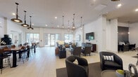 Located at 900 Doverside Dr in Apex, NC, this single-floor community clubhouse offers a thoughtfully designed gathering space that blends contemporary style with functional comfort. The open-concept layout is anchored by light-toned tile flooring throughout, creating a bright and cohesive atmosphere across all areas.

The main lounge is generously proportioned, featuring multiple distinct seating arrangements furnished with modern upholstered chairs and sofas in neutral tones, accented with pops of mustard yellow. Decorative pendant lights — including geometric wire and elongated teardrop styles — hang from elevated ceilings, adding visual interest. Large-format wall-mounted televisions are positioned throughout for easy viewing, complemented by curated abstract artwork on the walls.

Floor-to-ceiling windows and glass sliding doors flood the space with natural light while offering direct views of and access to the outdoor amenity areas. Several intimate seating nooks positioned near these openings provide semi-private spaces for smaller gatherings or quiet relaxation.

A well-appointed kitchen and dining area features dark cabinetry, stainless steel appliances, a dual high-top communal table with bar seating, open shelving, and dual wall-mounted screens — ideal for casual dining or community events. An additional bistro-style seating area near large exterior windows rounds out the space, offering a welcoming spot with views of the surrounding property.