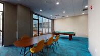 Located at 253 Academy St in Jersey City, NJ, this single-floor amenity space is designed to serve as a multifunctional social and recreational hub. The interior is anchored by vibrant teal patterned carpeting that runs throughout, creating a cohesive and energetic atmosphere across distinct activity zones.

The main room combines a game area and a gathering space, featuring a full-size pool table alongside a large wooden conference-style table surrounded by mustard yellow upholstered chairs. Recessed lighting in a drop tile ceiling provides consistent illumination throughout. Large floor-to-ceiling windows along one wall allow natural light to flow in while offering views of the adjacent outdoor patio.

A dedicated lounge nook includes a tan leather sectional sofa, two blue mid-century-style armchairs, and a wall-mounted flat-screen television, creating a comfortable media viewing area. The space also includes additional corridors with access to secondary rooms and utility areas.

The outdoor patio is finished with a stone paver surface and features a black dining table with vibrant red chairs, enclosed by low stone walls and fencing. The patio is accessible directly from the interior through the large glass doors, seamlessly connecting indoor and outdoor spaces.