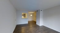 This single-story space showcases an open-concept layout that seamlessly connects the living area to the dining space and kitchen. The flooring throughout is dark hardwood-style, providing a rich, cohesive look that contrasts cleanly with the crisp white walls and white baseboards. A defining architectural feature is the pass-through window that opens from the kitchen into the living area, complete with a granite or stone-topped counter ledge — a practical and visually appealing detail that enhances the flow between spaces. The kitchen, visible through the pass-through, features cabinetry in a warm, neutral tone with a decorative tile backsplash, lending a touch of character to the cooking area. A drum-style pendant light fixture is suspended in the dining area, providing a focal point and warm ambient lighting. HVAC vents are integrated into the ceiling, ensuring year-round climate comfort. The layout is clean and functional, offering flexible furniture arrangement options. The overall ambiance is modern yet understated, with a neutral palette that provides an excellent foundation for a variety of interior design styles.