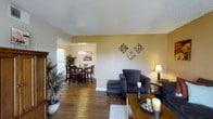 This single-story home offers a thoughtfully designed open-concept layout that seamlessly connects the living and dining areas, creating a sense of spaciousness and easy flow. The living area features rich hardwood flooring that extends throughout the main space, complemented by warm, neutral-toned walls that lend a welcoming ambiance. A vaulted ceiling adds vertical dimension to the room, enhancing the overall sense of openness. Decorative diamond-shaped wall art adds a stylish focal point to the living space, while a floor lamp provides warm accent lighting. The adjacent dining area is visible through a wide open passage, complete with a dark wood dining set and an elegant chandelier overhead. Natural light filters in through a nearby window, brightening the dining space and highlighting the greenery of potted plants that add life to the room. The textured ceiling treatment is consistent throughout, adding subtle architectural character. Overall, the space presents a cohesive, comfortable atmosphere with a balance of natural materials, warm lighting, and decorative touches that make it feel both functional and inviting.