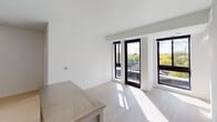 This contemporary single-floor unit showcases a clean, minimalist design defined by crisp white walls, light hardwood flooring, and an abundance of natural light throughout. The open-concept kitchen is a functional highlight, featuring white upper and lower cabinetry, dark countertops with a subtle tile backsplash, and a full suite of stainless steel appliances including a refrigerator, electric range, microwave, and dishwasher. A central island with a stainless steel countertop provides additional prep space and casual seating. The living area is bright and airy, with floor-to-ceiling black-framed windows and a glass door opening onto a private balcony, offering pleasant views of surrounding greenery. The bedroom mirrors this design sensibility, featuring the same light flooring and large black-framed windows that draw in generous daylight and frame outdoor views. The full bathroom is finished in a modern style, with light wood-look flooring, a full-size soaking tub with a chrome shower rod and fixtures, a wall-mounted mirror, and a floating vanity with ample under-cabinet storage and an integrated white sink. Throughout the unit, the consistent use of neutral tones, clean lines, and quality materials creates a cohesive and inviting living environment.