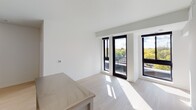 This contemporary single-floor unit showcases a clean, minimalist design defined by crisp white walls, light hardwood flooring, and an abundance of natural light throughout. The open-concept kitchen is a functional highlight, featuring white upper and lower cabinetry, dark countertops with a subtle tile backsplash, and a full suite of stainless steel appliances including a refrigerator, electric range, microwave, and dishwasher. A central island with a stainless steel countertop provides additional prep space and casual seating. The living area is bright and airy, with floor-to-ceiling black-framed windows and a glass door opening onto a private balcony, offering pleasant views of surrounding greenery. The bedroom mirrors this design sensibility, featuring the same light flooring and large black-framed windows that draw in generous daylight and frame outdoor views. The full bathroom is finished in a modern style, with light wood-look flooring, a full-size soaking tub with a chrome shower rod and fixtures, a wall-mounted mirror, and a floating vanity with ample under-cabinet storage and an integrated white sink. Throughout the unit, the consistent use of neutral tones, clean lines, and quality materials creates a cohesive and inviting living environment.