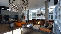 This contemporary lounge space makes an immediate impression with its bold industrial-modern design aesthetic. The centerpiece of the room is a spectacular cascading chandelier composed of dozens of exposed Edison-style bulbs suspended at varying heights, casting a warm, dynamic glow throughout the space. Soaring ceilings and floor-to-ceiling glass walls flood the interior with abundant natural light while offering views of the surrounding urban environment. Structural cylindrical concrete columns punctuate the open floor plan, reinforcing the industrial character of the space. A curated large-scale motorcycle artwork mounted on a textured stone panel serves as a dramatic focal point, adding an edgy, creative dimension to the interior. The seating arrangement is thoughtfully composed, featuring a mix of curved orange sofas, leather club chairs in warm cognac tones, and sleek grey upholstered pieces that invite relaxed conversation and social gathering. A modern fireplace anchors one wall, contributing warmth and ambiance. Polished hardwood flooring grounds the space, contrasting beautifully with the raw concrete and metal finishes. The overall atmosphere is energetic yet comfortable, blending artistic flair with functional design to create a compelling and memorable gathering space.