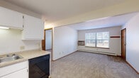 This single-floor unit offers a practical and well-organized layout designed for comfortable everyday living. The kitchen is a standout feature, configured in an L-shape with ample white cabinetry providing generous storage, light-toned laminate countertops, and tile flooring. It is fully equipped with a white electric range, built-in microwave, refrigerator, and black dishwasher, all illuminated by overhead fluorescent lighting. The kitchen opens directly into the living area, which is carpeted and benefits from a large window that invites natural light and views of the outdoors. A wall-mounted air conditioning unit and baseboard heating ensure year-round climate comfort throughout the space. The bedroom is a well-proportioned room featuring plush carpet, a ceiling fan with light fixture, a window overlooking the exterior, and baseboard heating. An additional room provides flexible space suitable for use as a secondary bedroom or office. Storage is addressed by a dedicated walk-in closet outfitted with multiple wood shelving units and hanging rods. The bathroom features tile flooring, a full-size tub and shower combo, a vanity with sink, and a toilet. Warm wood-toned trim and doors are consistent throughout, contributing to a cohesive, neutral aesthetic across the entire unit.